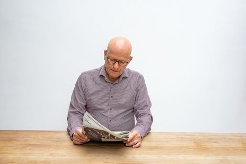 Free Elderly man sitting at a wooden table reading a newspaper indoors. Stock Photo