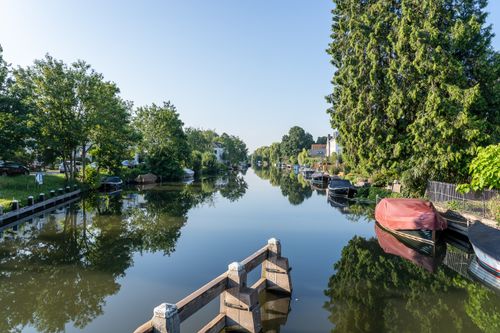 Free Peaceful canal lined with boats and lush trees reflecting in the water under a clear summer sky. Stock Photo