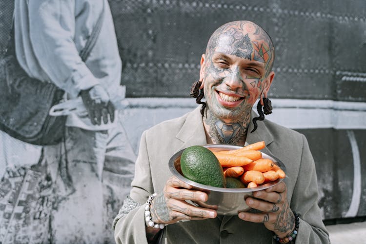 A Tattooed Man Holding Steel Bowl With Fresh Vegetable And Fruit