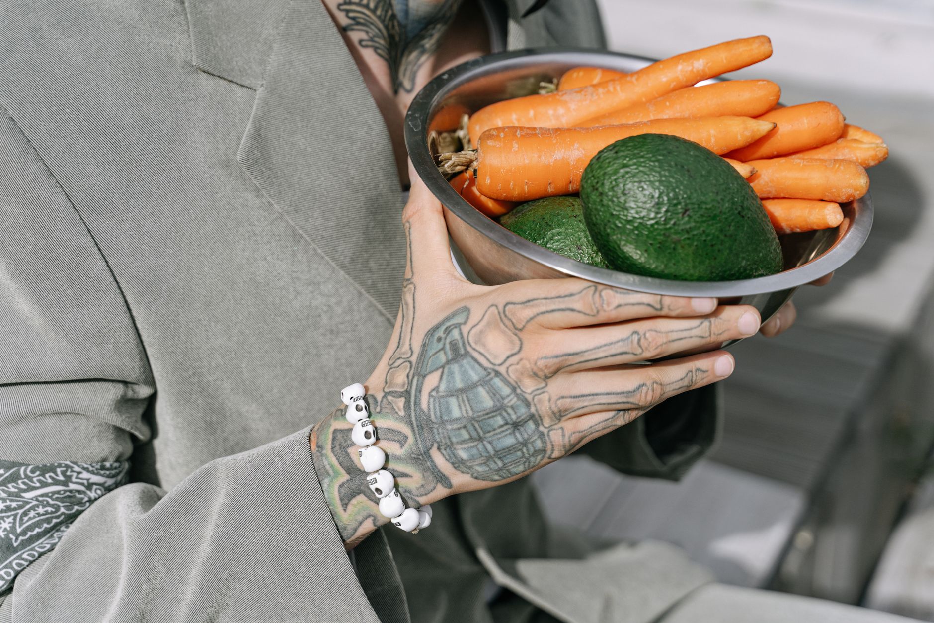 A person with tattoos holds a bowl of fresh avocados and carrots, showcasing healthy living.