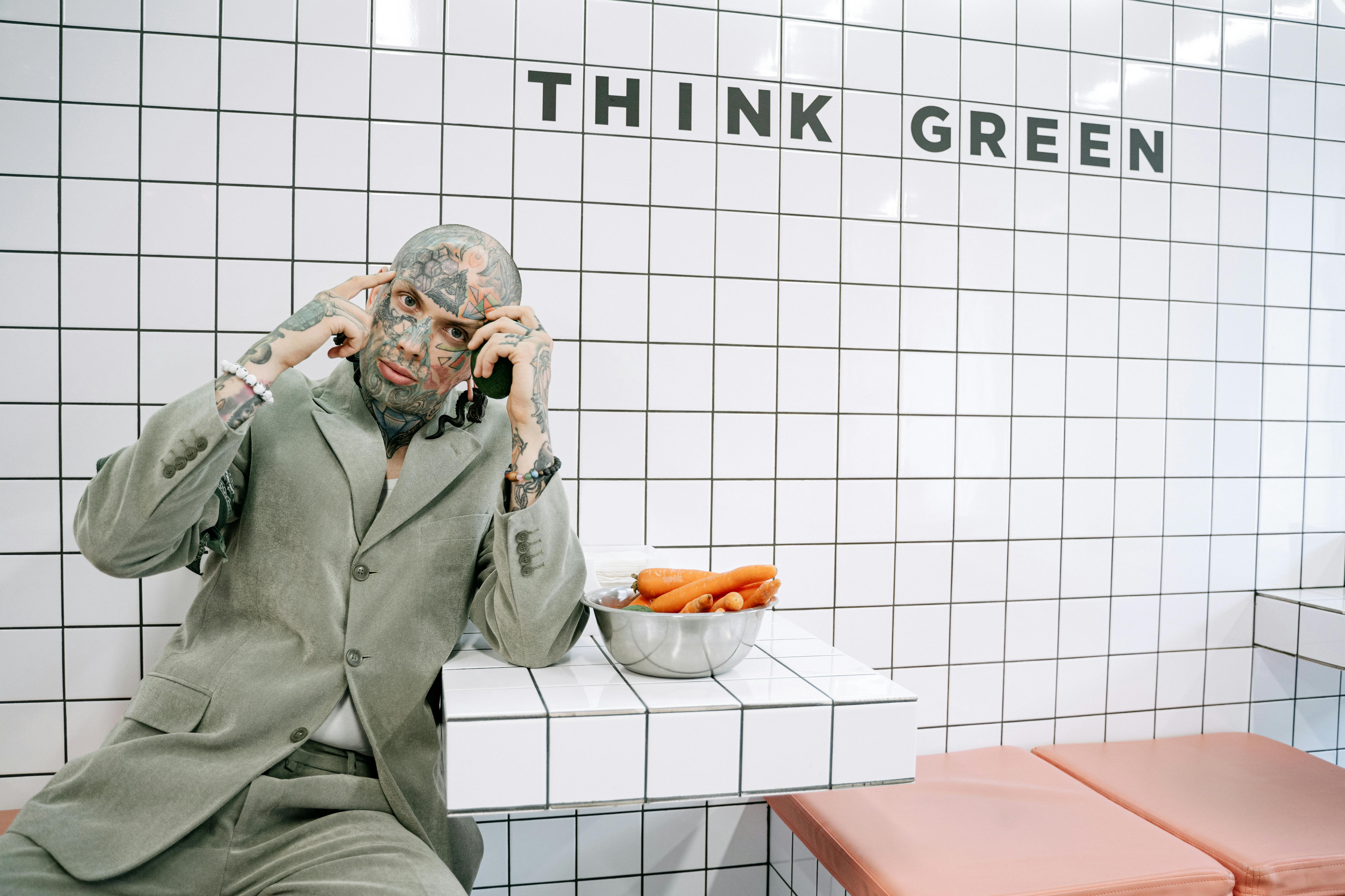 A tattooed man in a suit sits beside a bowl of carrots on a tiled bench with 'Think Green' text.