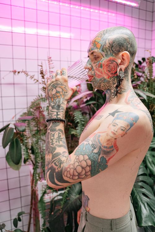 Free Shirtless tattooed man drinking water in a modern indoor setting with plants. Stock Photo