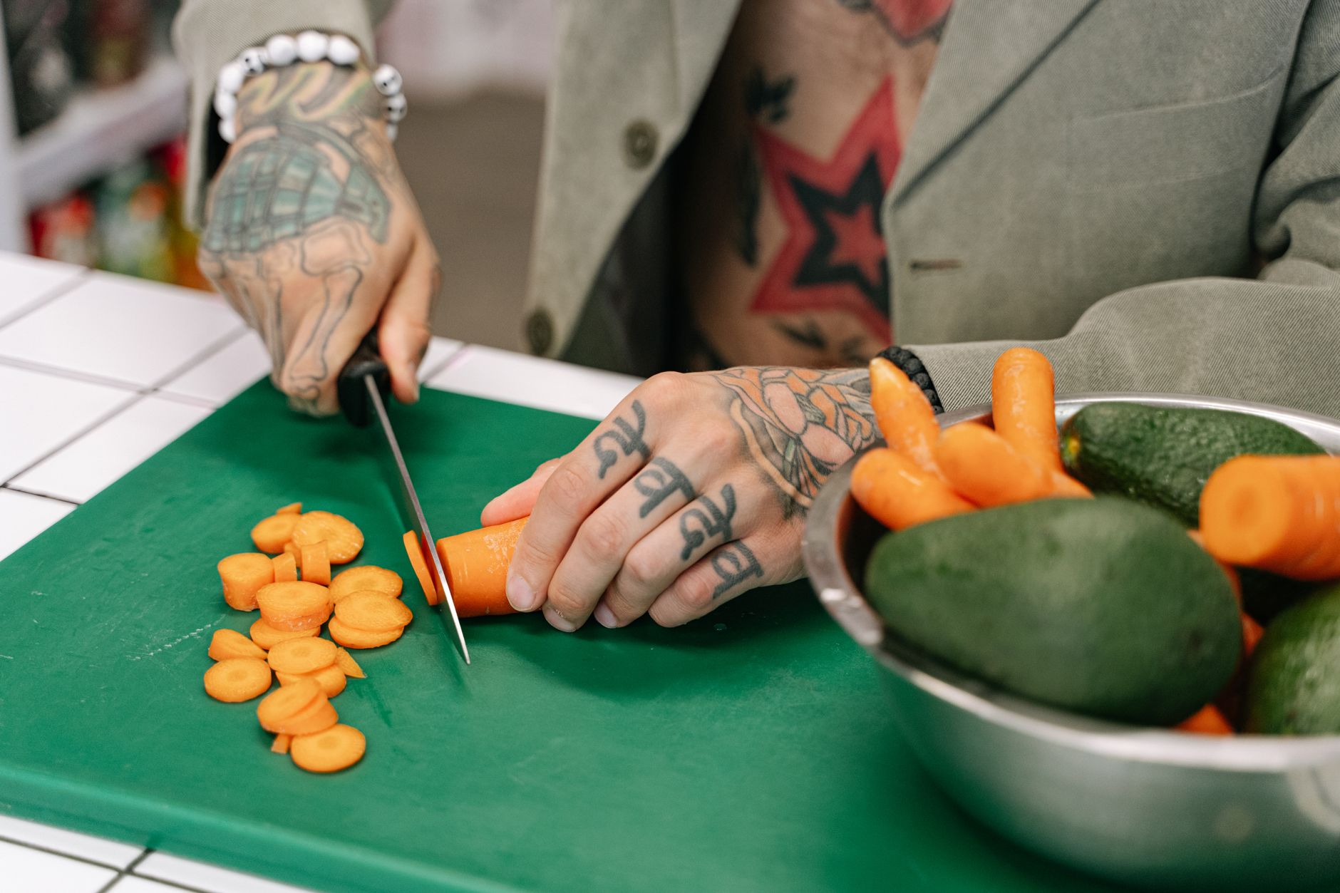 A tattooed person cutting carrots on a green chopping board with avocados nearby, showcasing culinary skill and style.