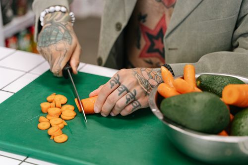 Free A tattooed person cutting carrots on a green chopping board with avocados nearby, showcasing culinary skill and style. Stock Photo