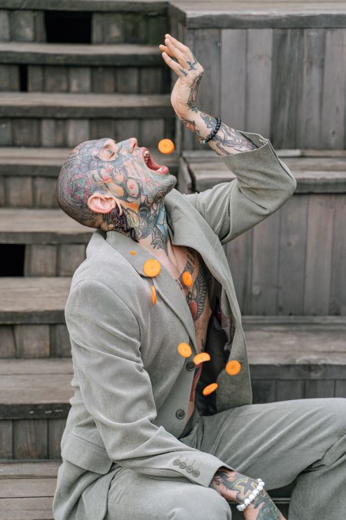 Free A tattooed man in a suit joyfully tosses sliced carrots in the air while sitting outdoors on stairs. Stock Photo
