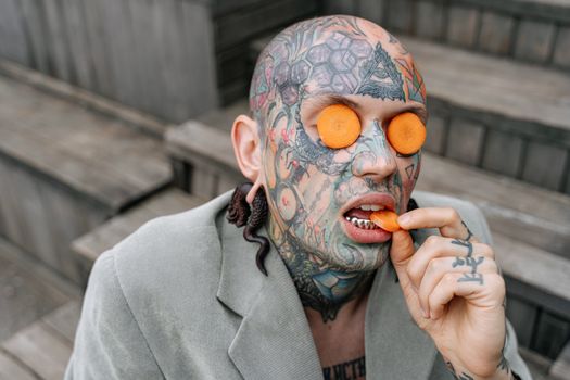 一名秃头男子，脸上和手上有鲜艳的纹身，在户外吃着胡萝卜片
