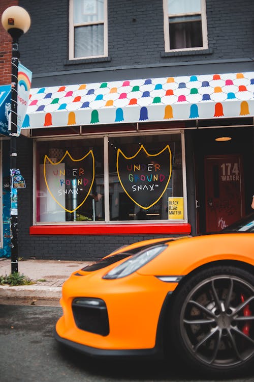 Orange modern sports car driving on empty city street against residential building with shop in daytime