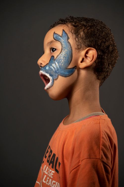 Free Young boy in orange shirt with blue face paint and open mouth on dark background. Stock Photo