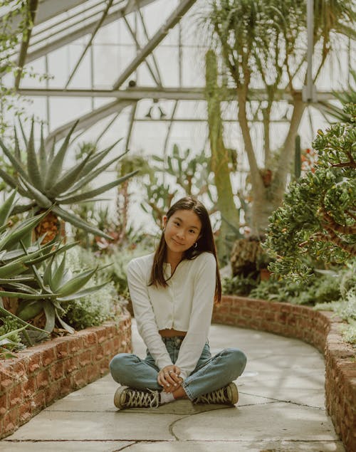 Full body of positive ethnic female teenager in stylish outfit sitting with crossed legs on pathway among exotic plants in greenhouse and smiling
