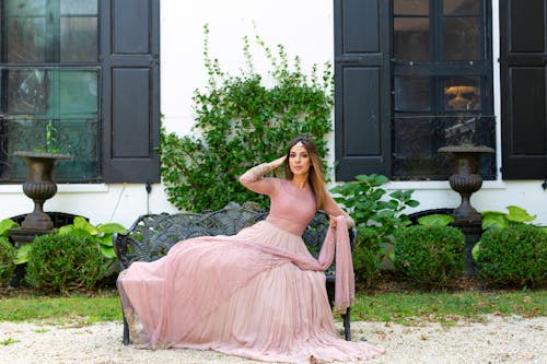 Free Full body of ethnic female wearing pink wedding dress with long hem and Maang Tikka looking at camera and touching hair while sitting on metal bench on terrace near residential building Stock Photo