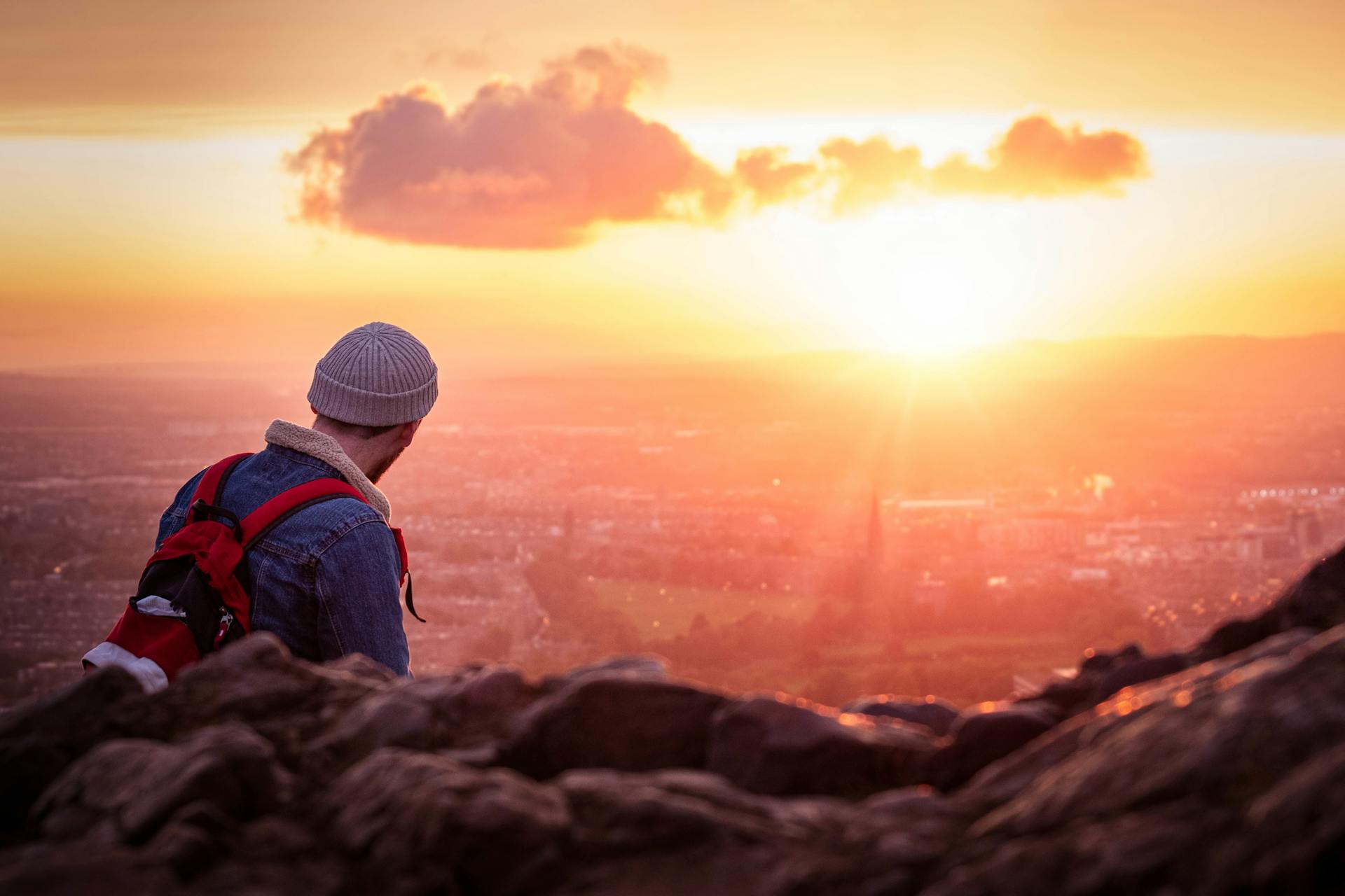 Backpacker overlooking Edinburgh cityscape at sunset from hilltop.