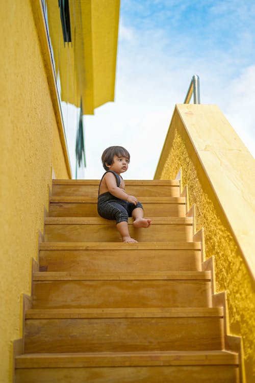 Free From below of cute barefoot child sitting on stairs under cloudy sky while looking away Stock Photo