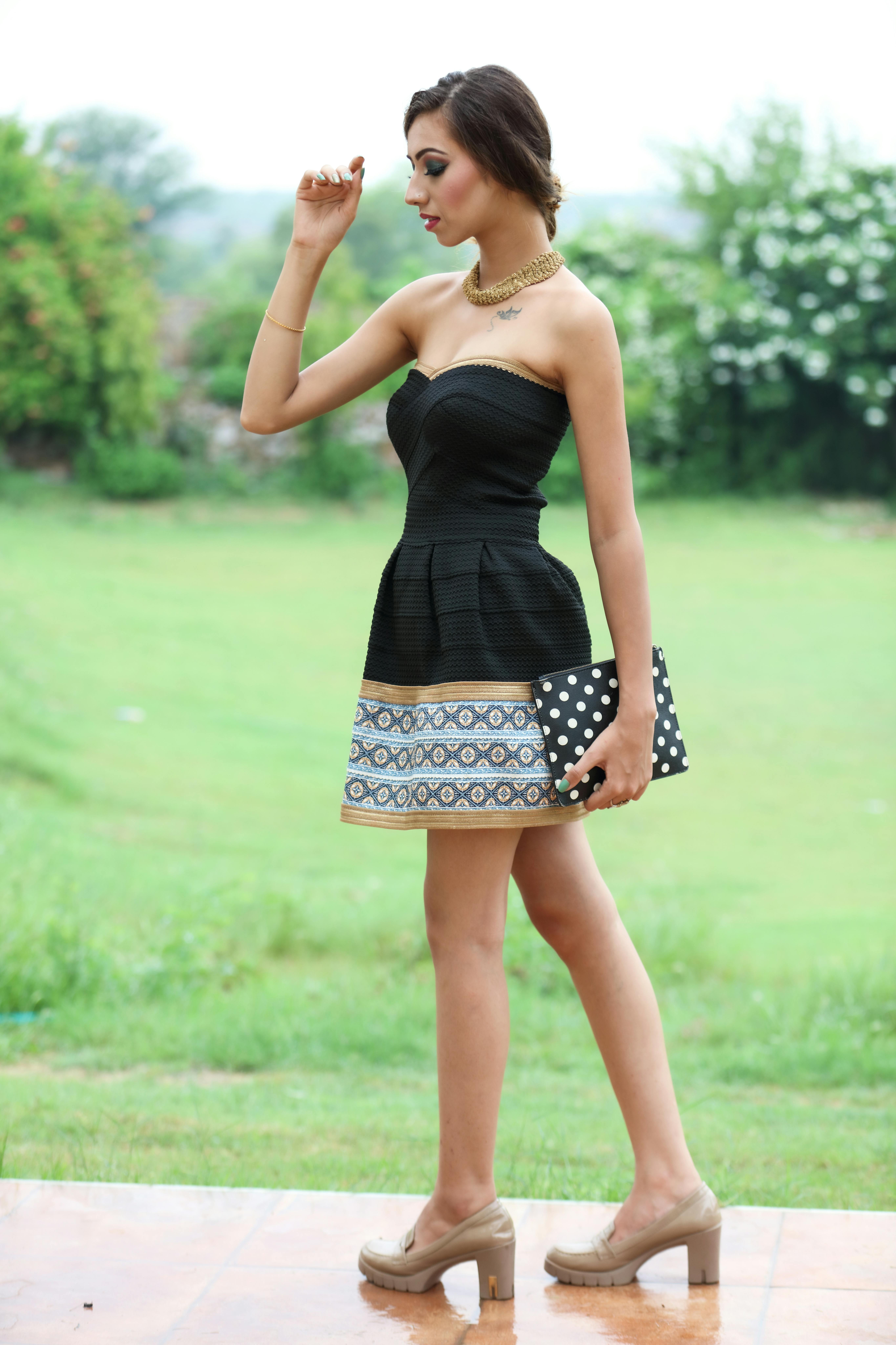Side view full length of serious young slim female in mini dress standing on veranda with purse near green grassy meadow and bushes with plants in summer day