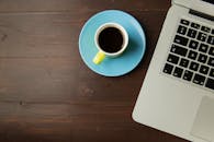 咖啡杯放在茶托上，顶视图旁边是木桌上的笔记本电脑，非常适合在家工作的主题