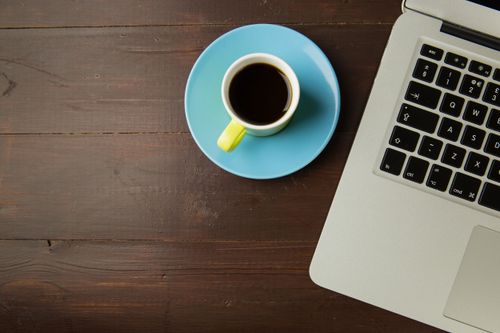 Free A top view of a coffee cup on a saucer next to a laptop on a wooden desk, ideal for work-from-home themes. Stock Photo