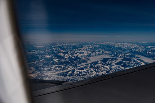 View of tall bare rocks with snow on tops under blue sky with clouds from window of aircraft