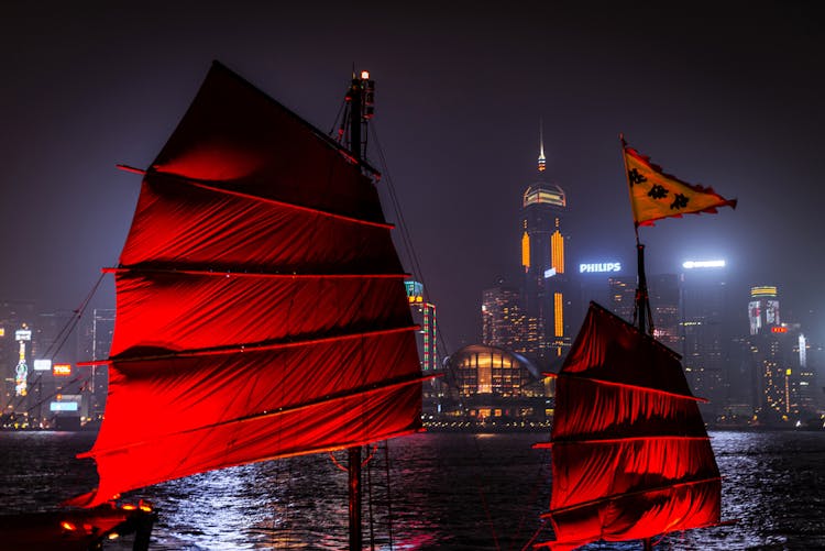 Red Sail Boat On Body Of Water Near City Buildings During Night Time