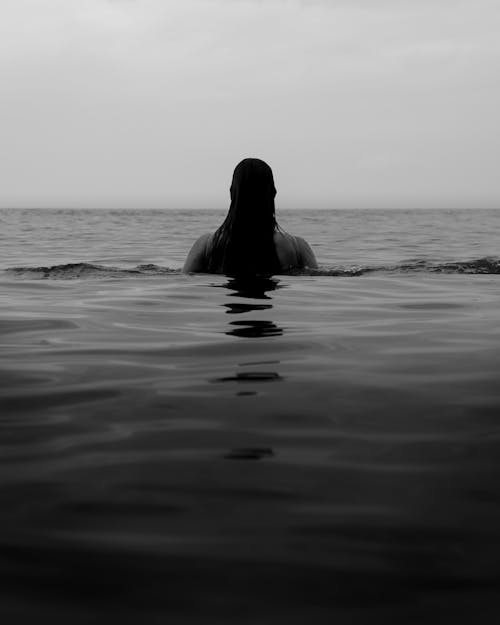 Black and white of anonymous dark haired female swimming in rippling sea against skyline