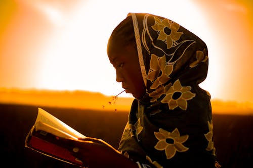 Side view of black female in traditional wear reading book while standing against bright skyline