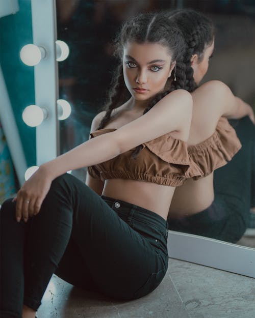 Charming female with braids and trendy makeup leaning on mirror with lamps and looking at camera