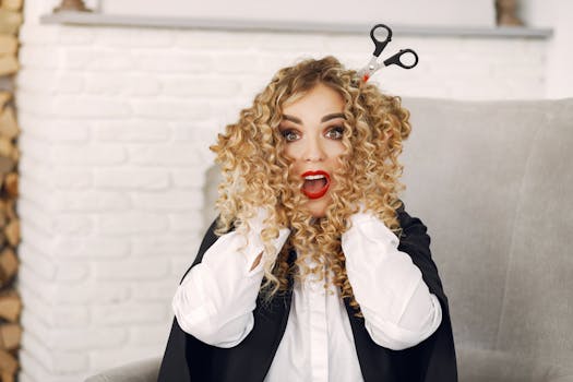 A woman with curly hair and surprised expression, sitting indoors with scissors in her hair.