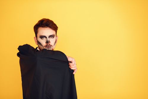 Free Man in skull makeup and black cape on yellow background. Halloween. Stock Photo