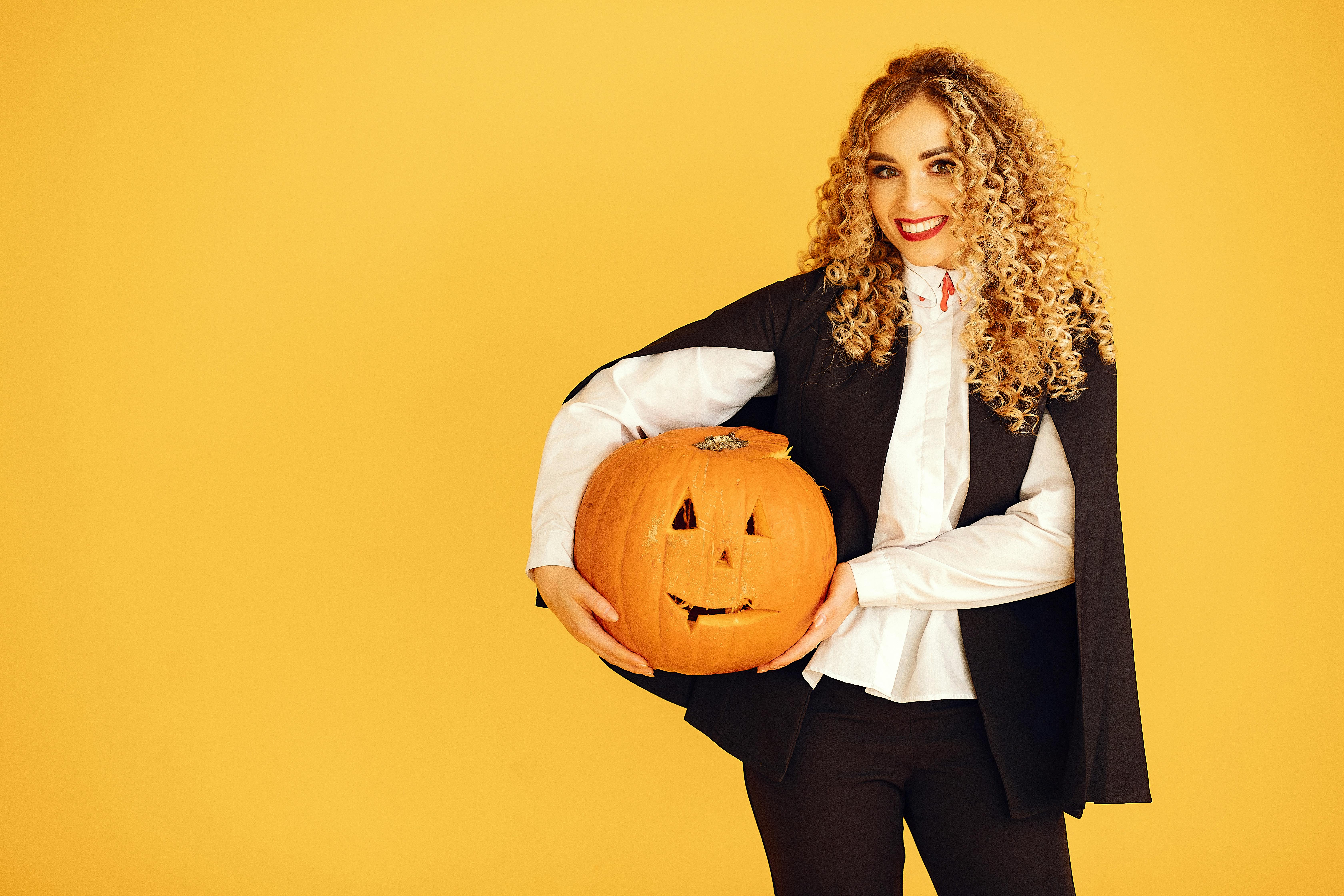 一位身穿德古拉服装的年轻女子面带微笑，手拿南瓜，背景是充满活力的黄色
