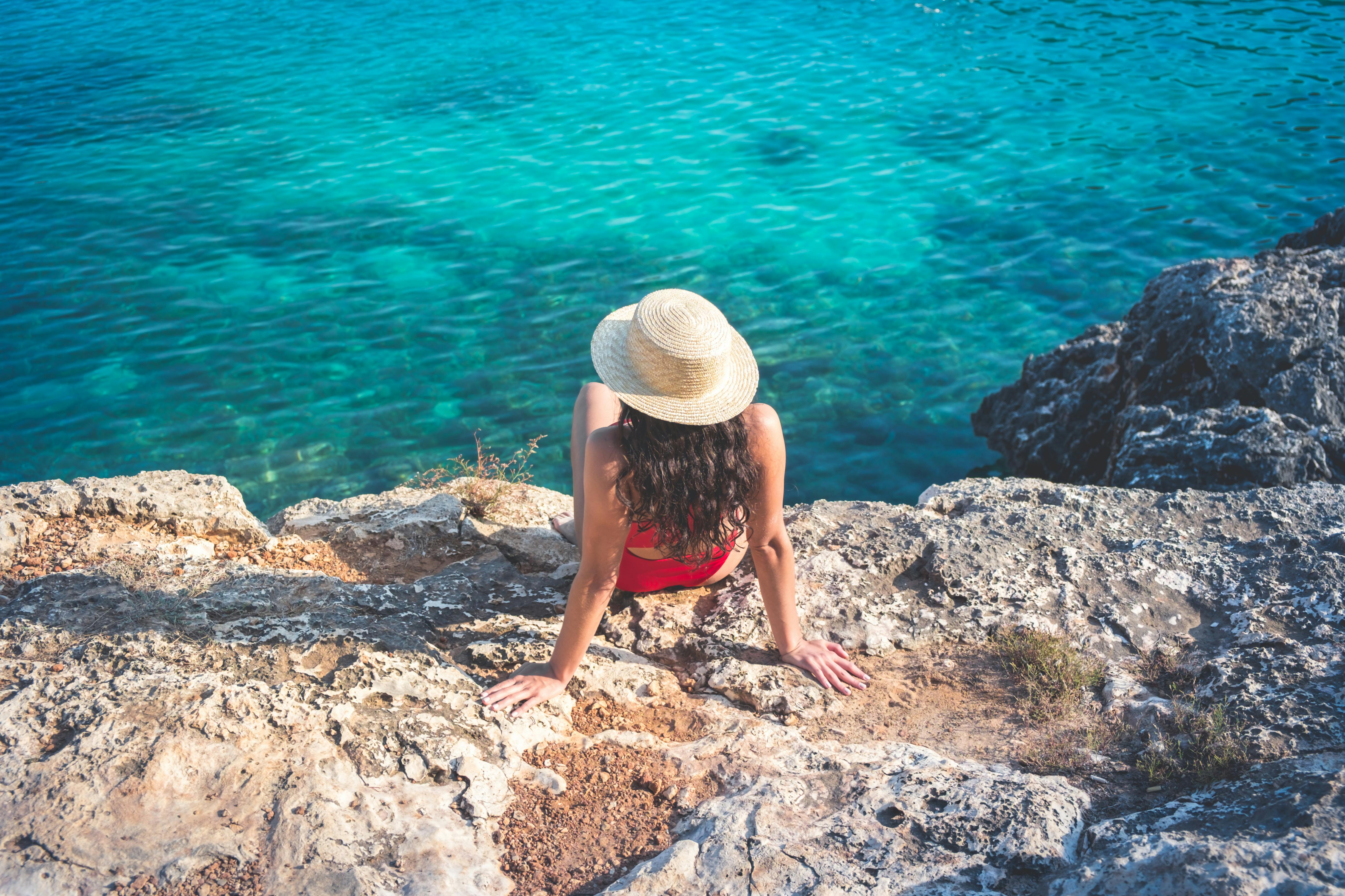 Free A woman in a red bikini and beach hat enjoys the summer sun on rocky cliffs by clear blue ocean waters. Stock Photo