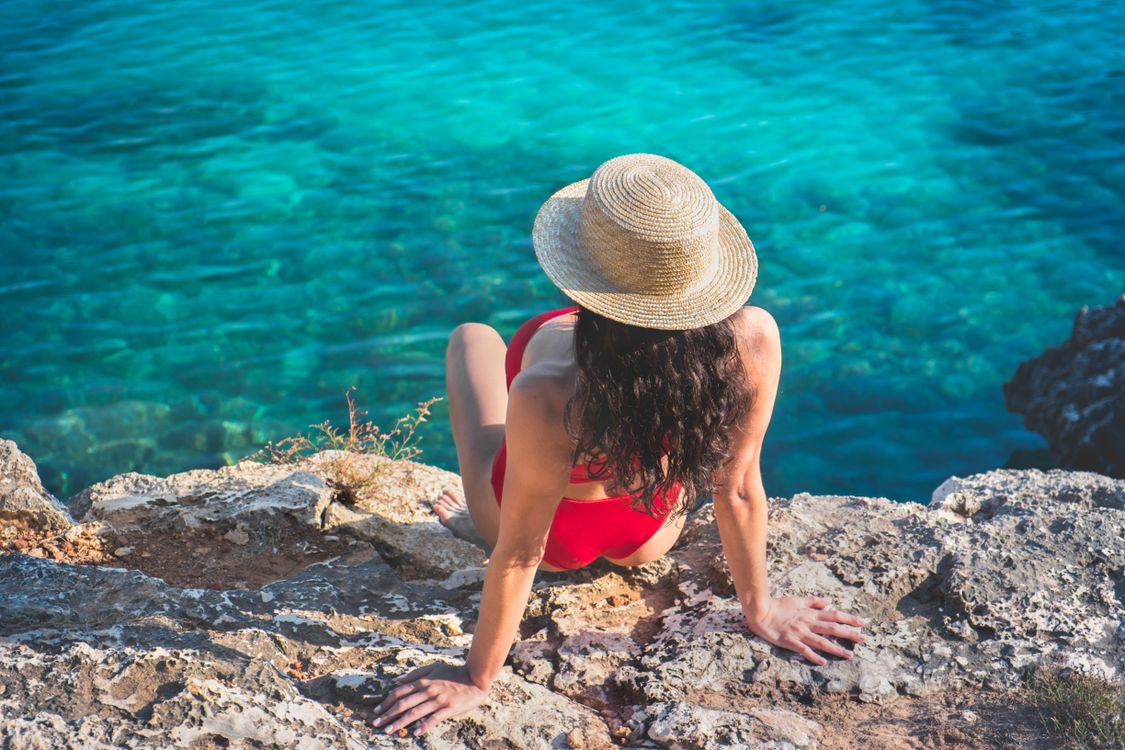 Free A woman in a red swimsuit and straw hat relaxes on a rocky beach overlooking the ocean. Stock Photo