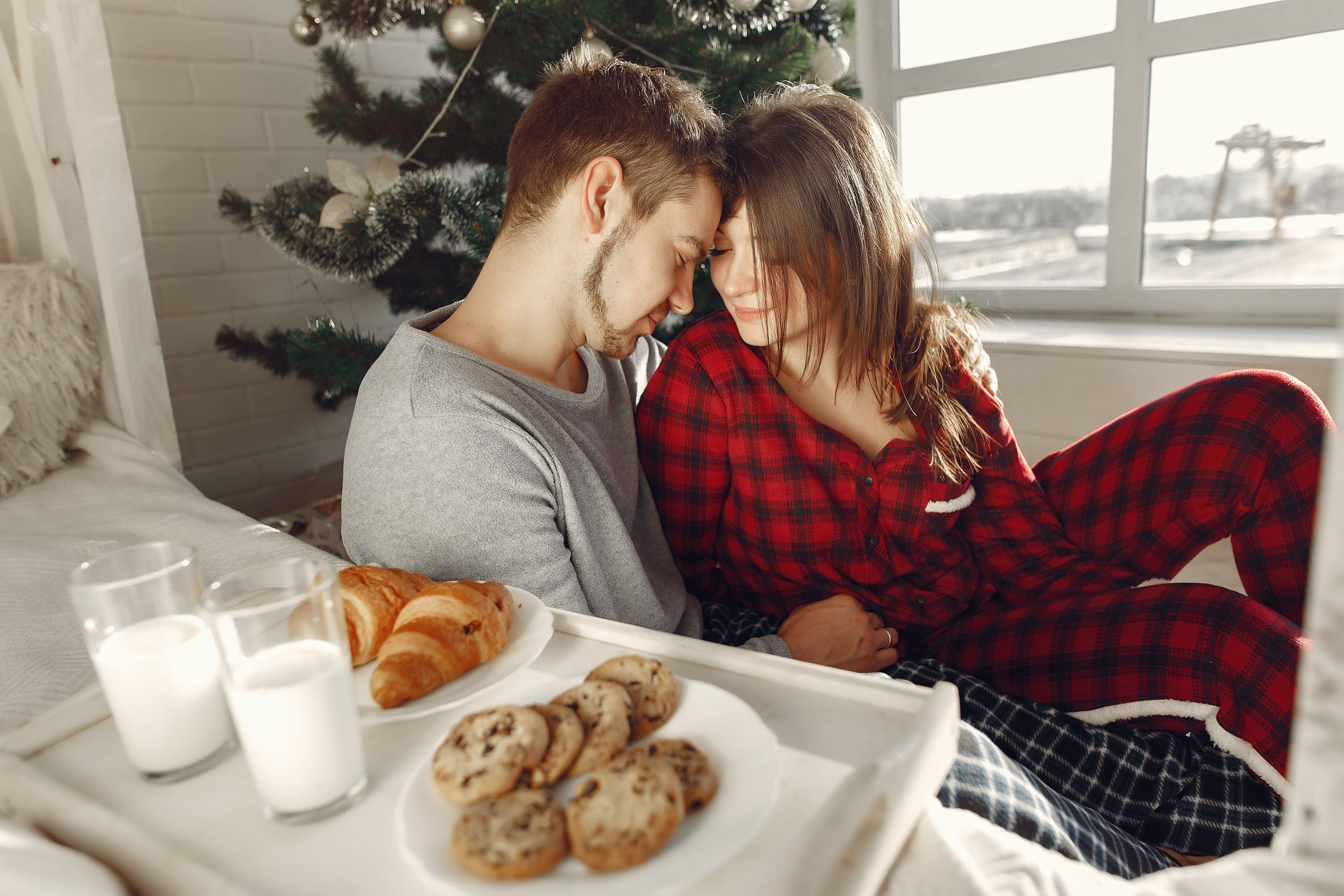 A couple in pajamas enjoys breakfast by a Christmas tree in a warm, festive setting.