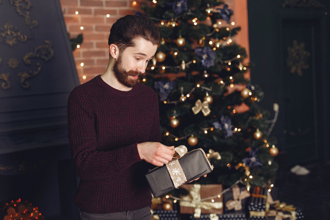 Man in a burgundy sweater opening a wrapped gift box beside a lit Christmas tree