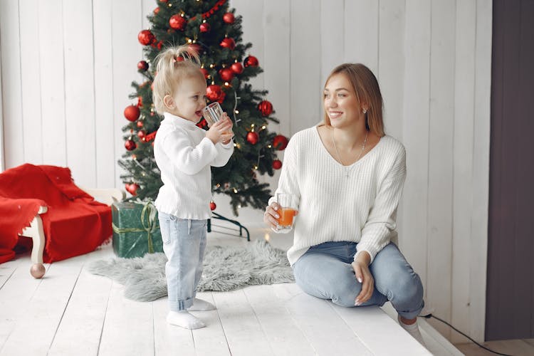 Mom Sitting By Christmas Tree And Drinking Juice With Playing Daughter