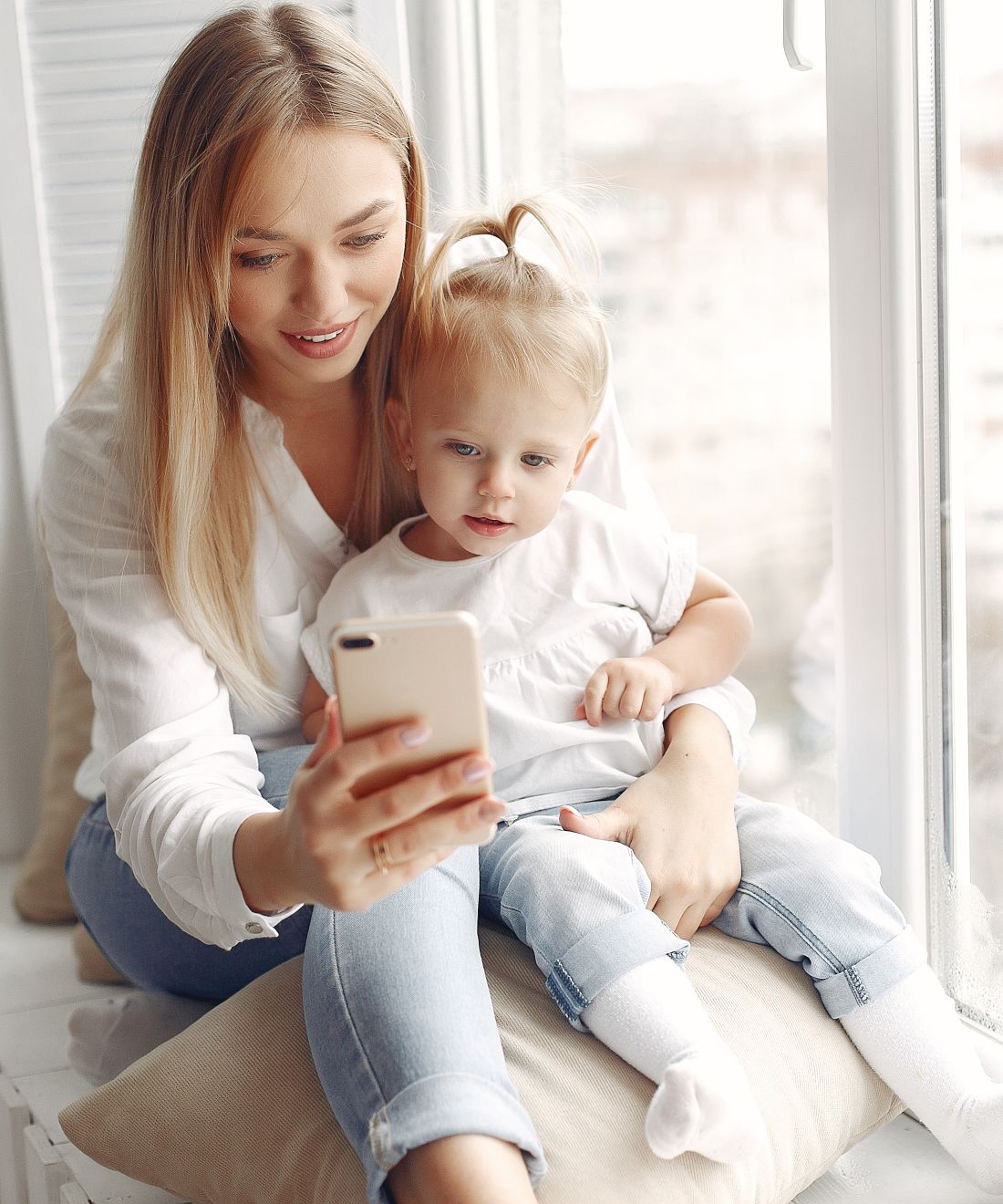 Una mamma bionda e la sua bambina guardano insieme lo smartphone davanti alla finestra di casa