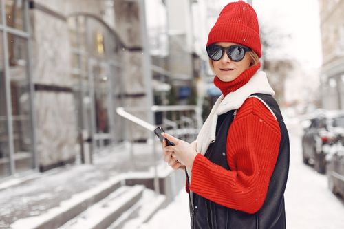 Free Stylish woman wearing winter clothes and sunglasses on a snowy city street, holding a cellphone. Stock Photo