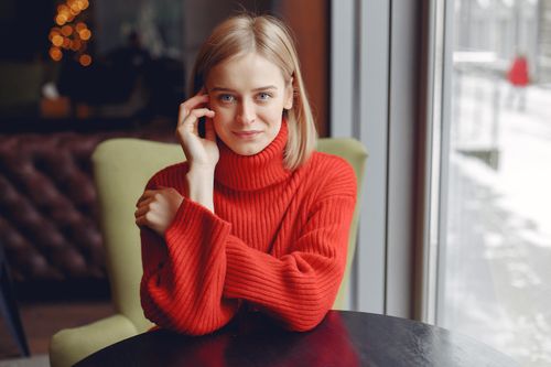 Free A woman sits by a window, wearing a red knitted sweater, with a calm expression. Stock Photo