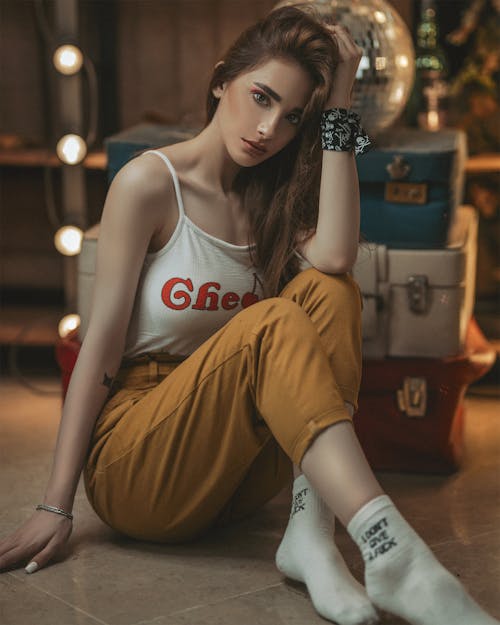 Full body of young slim female with gentle makeup in trendy outfit sitting on floor near vintage suitcases and disco ball