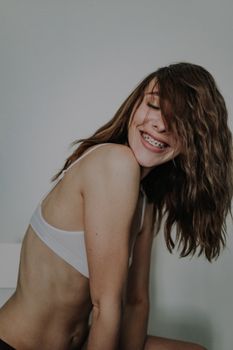 侧面靠墙的快乐年轻女性，身穿白色胸罩，双手倚着白墙，闭眼微笑露出牙套