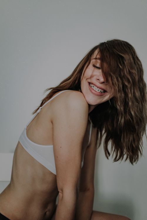 Free Side view happy young female wearing white bra leaning on hands against white wall and smiling with eyes closed showing teeth braces Stock Photo