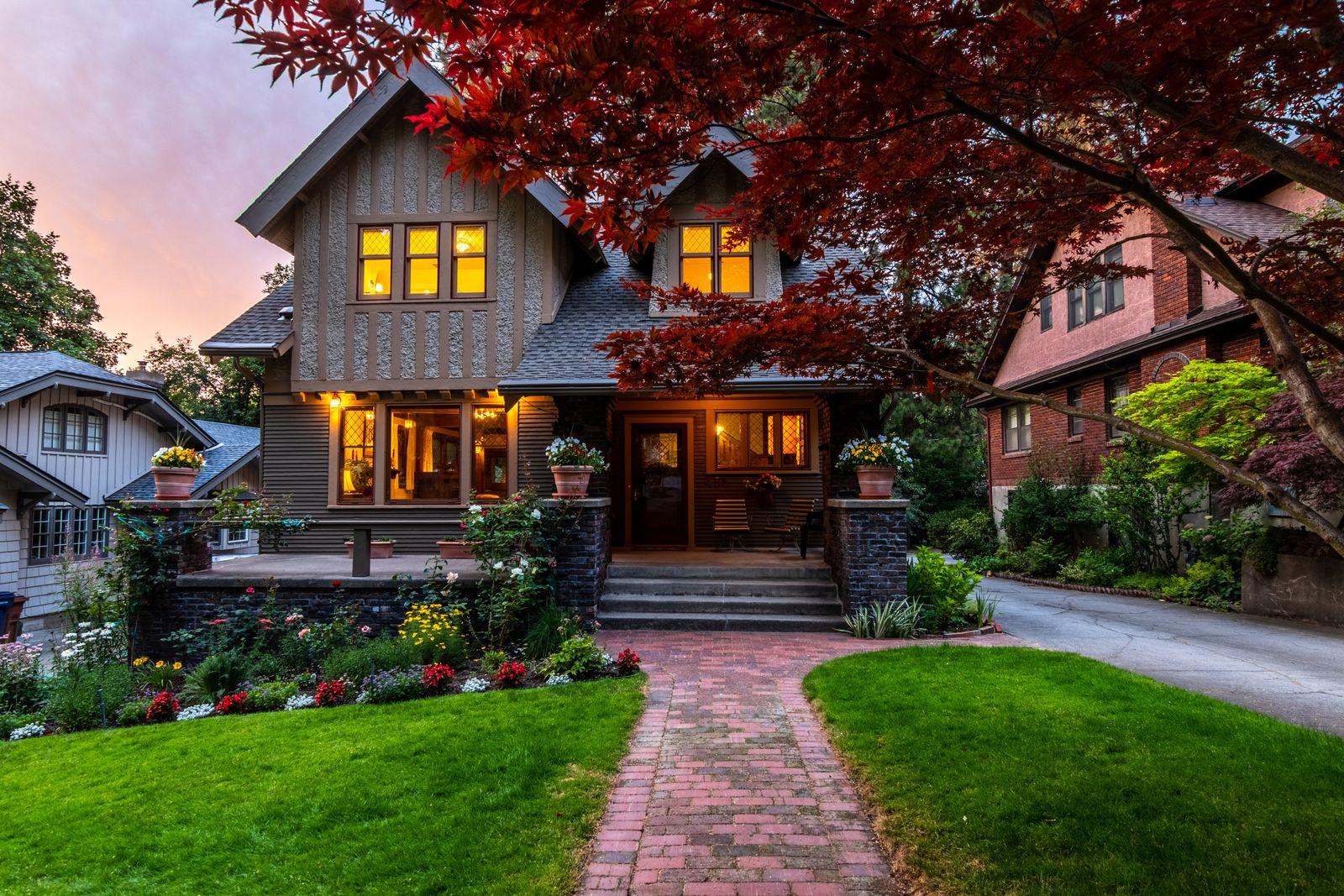 Two-story Craftsman-style home with stucco-and-timber gables and lit windows at dusk, a brick paver walkway crossing a manicured lawn and flower beds, West Highlands, Naperville IL