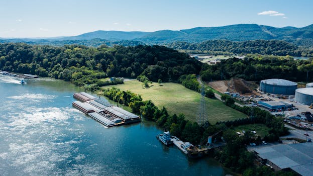 Picturesque drone view of rippling river flowing among lush green trees near industrial area in mountainous valley on sunny day