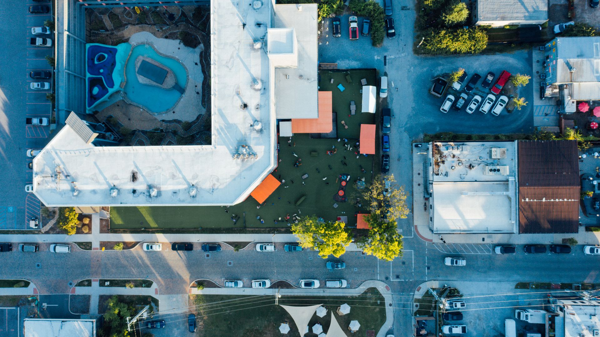 Aerial view of an urban area showing buildings, a pool, and parked cars in daylight.