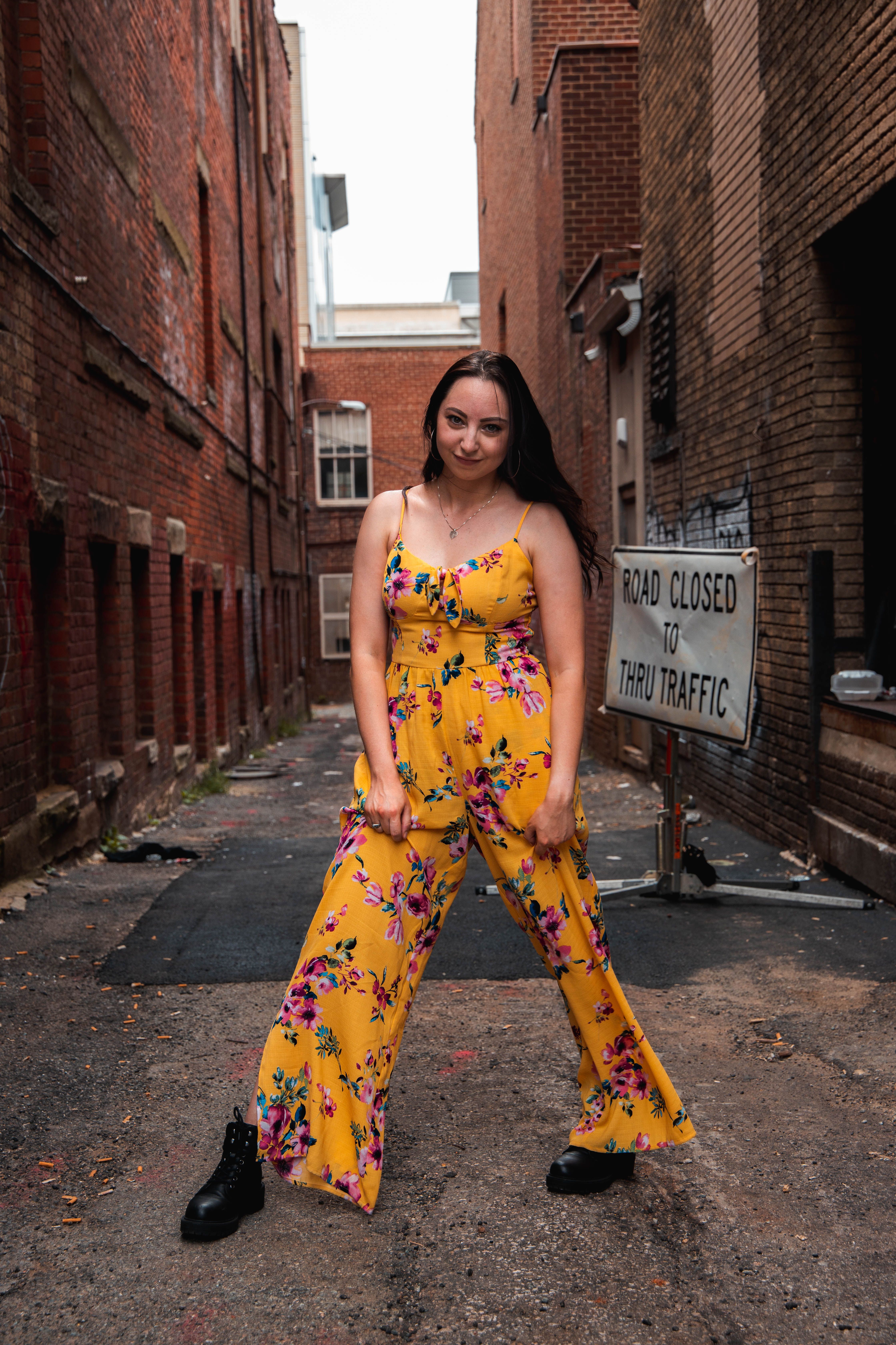 A woman in a vibrant floral dress poses confidently in an urban alley with brick walls.
