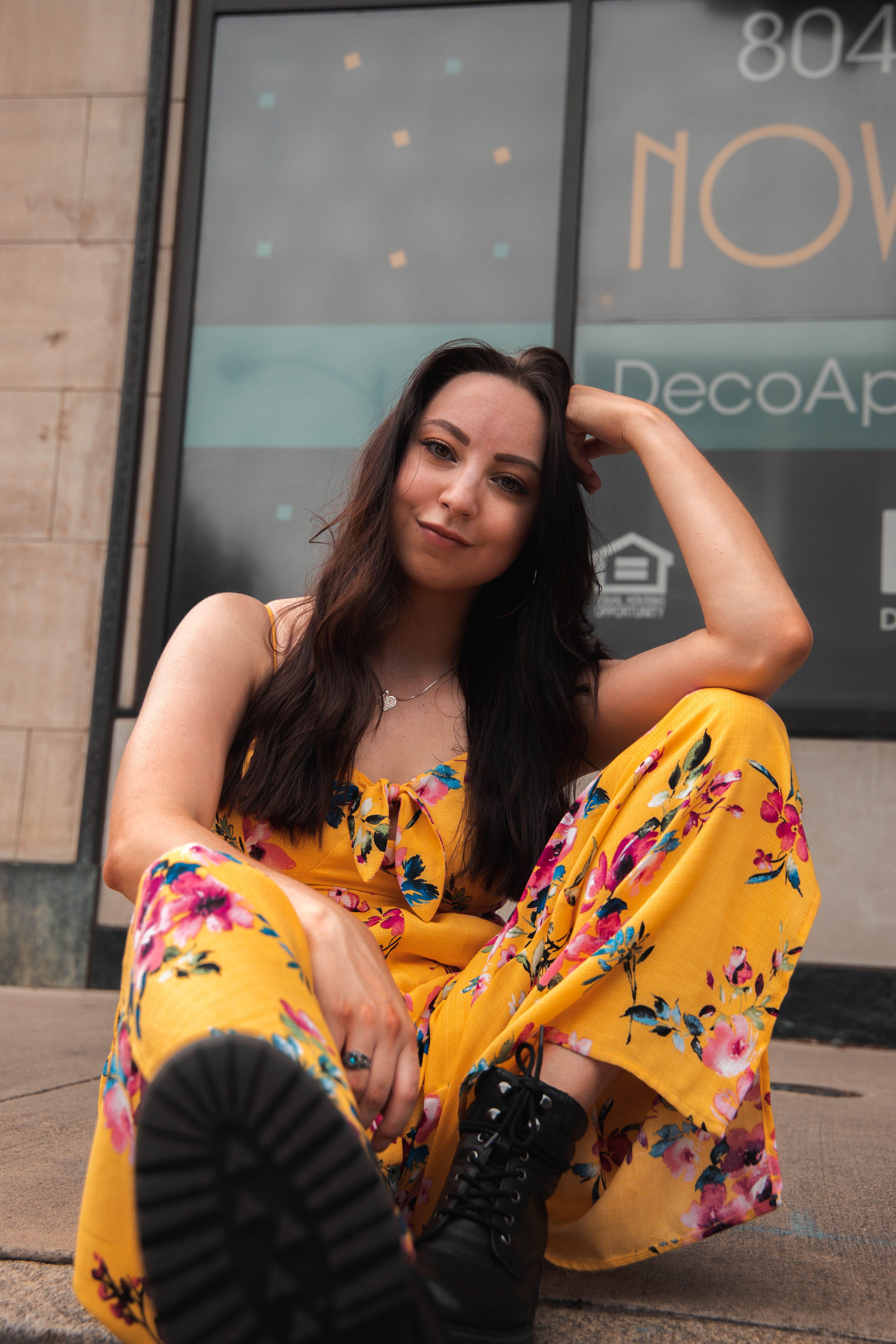 A woman in a vibrant floral dress posing outdoors by a glass window.
