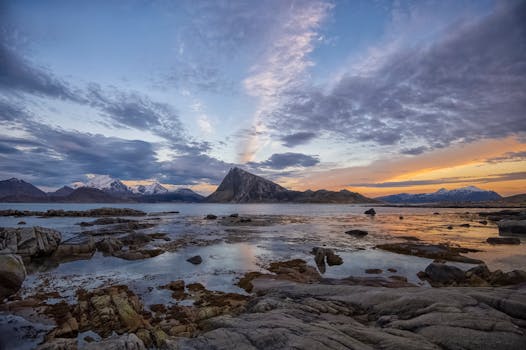 Picturesque scenery of rocky frozen shore of sea near snowy mountains against cloudy sunset sky in Lofoten archipelago