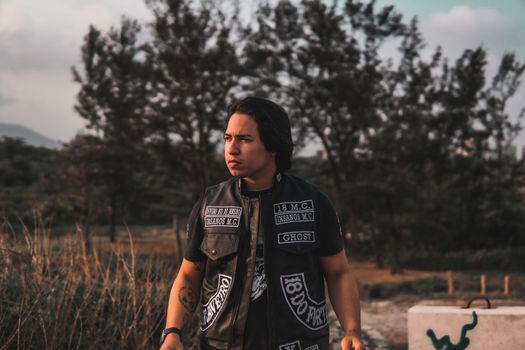 Man wearing leather vest with tattoos, set against a natural outdoor background during the day.