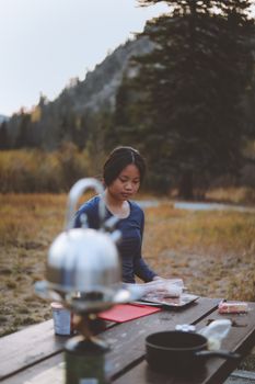 Woman camping and preparing food outdoors in a serene forest setting during the day.