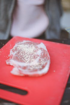 Raw meat sprinkled with salt on a red cutting board in a kitchen setting.