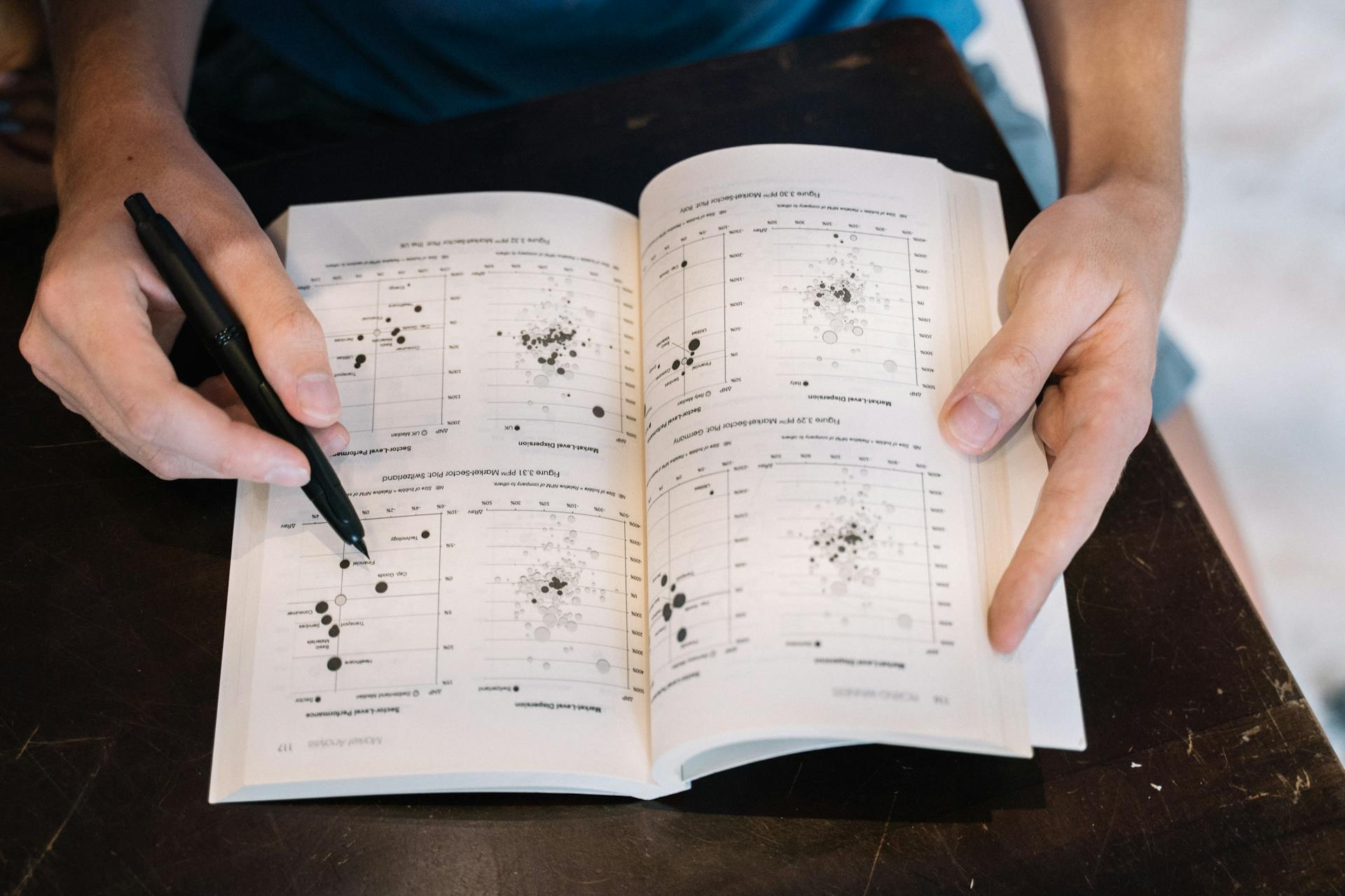 Person analyzing data charts in a book using a pen, close-up perspective.