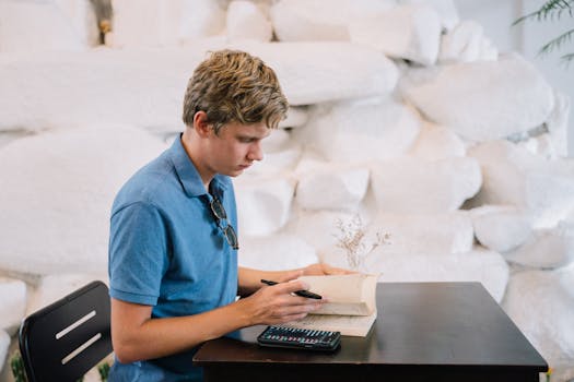 A young adult male in a blue polo shirt studies at a table indoors, using a book and smartphone.