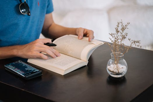 A man studying indoors with a book, smartphone, and dried plant on a table.